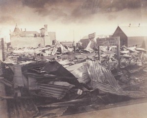 Rubble of buildings destroyed by fire, showing stack of corrugated iron and other rubble, sign still standing reads: Bennett's Pharmacy / Temporary Premises / next / Romney Studios. Man standing among rubble in background.